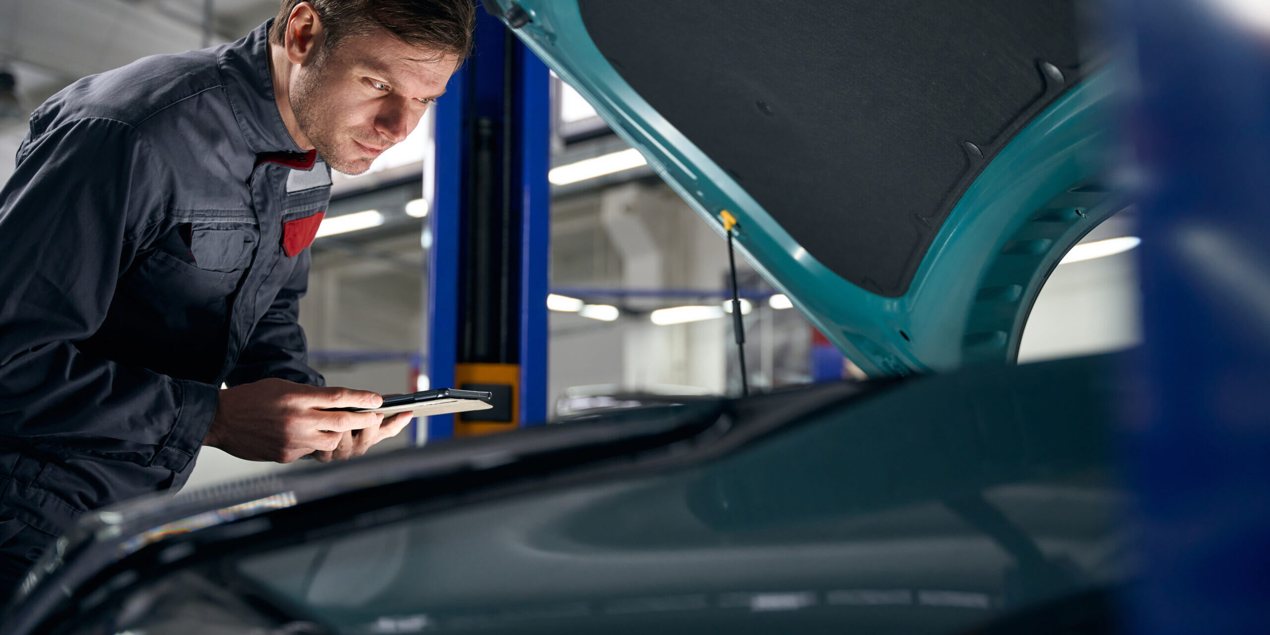 Male in protective clothes standing in tire fitting near car with open hood and holding laptop